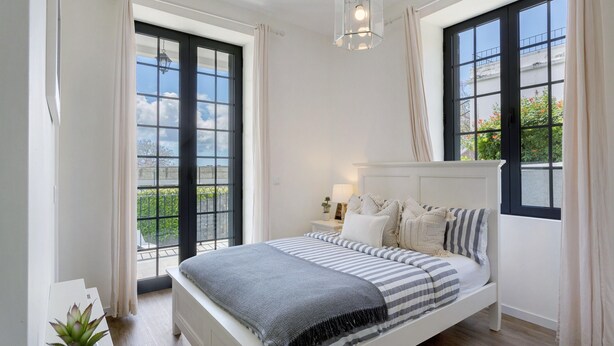 Un dormitorio con cama, mesita de noche, lámpara y ventanas amplias.