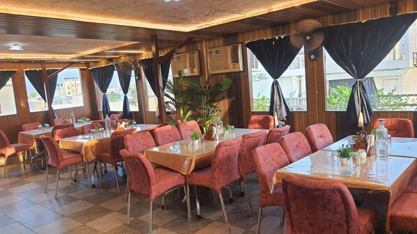 Un restaurante con mesas y sillas, ventanas amplias con cortinas y unidades de aire acondicionado.