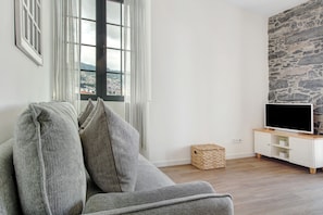 Una sala de estar con un sofá, un televisor sobre un mueble de madera, una pared de piedra y una ventana con cortinas.