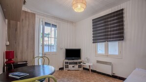 A room with a television on a wooden stand, a dining table with chairs, a radiator, and a window with blinds. - Studio Apartment 'Chez Lina -  Avec Terrasse' with Private Terrace and Wi-Fi (Le Lavandou)