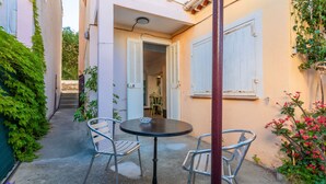 A narrow alleyway with a small patio area featuring a round table and two chairs, a door leading to an interior space, and a wall with climbing plants. - Studio Apartment 'Chez Lina -  Avec Terrasse' with Private Terrace and Wi-Fi (Le Lavandou)