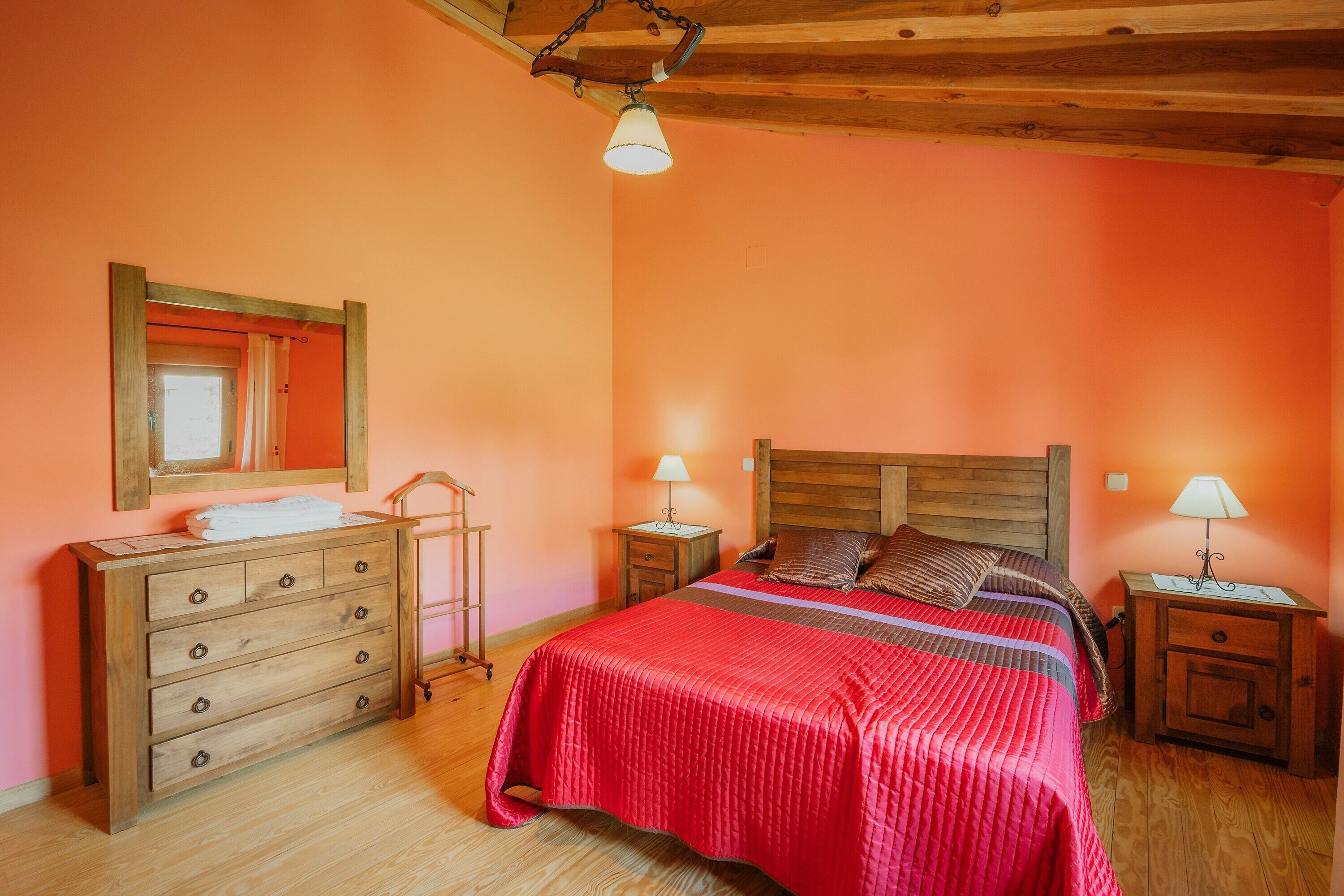 Une chambre avec une commode en bois, un lit recouvert d’une couverture rouge, deux tables de chevet avec des lampes et un plafond en bois.