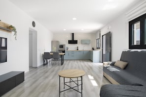A modern living room with a sofa, a coffee table, and a TV. There is a kitchen area with cabinets and a microwave.