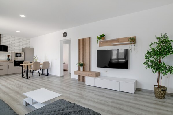A modern living space with a dining area, a TV mounted on a wooden panel, a large potted plant, and a kitchen in the background.