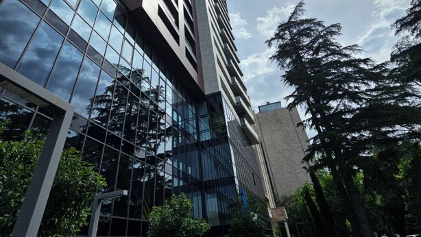 Un immeuble moderne de grande hauteur, doté de fenêtres en verre réfléchissant, entouré de verdure et d’autres bâtiments.