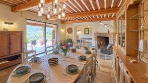 A dining area with a long table set for six, wooden beams on the ceiling, a fireplace, and a view of the outdoors.