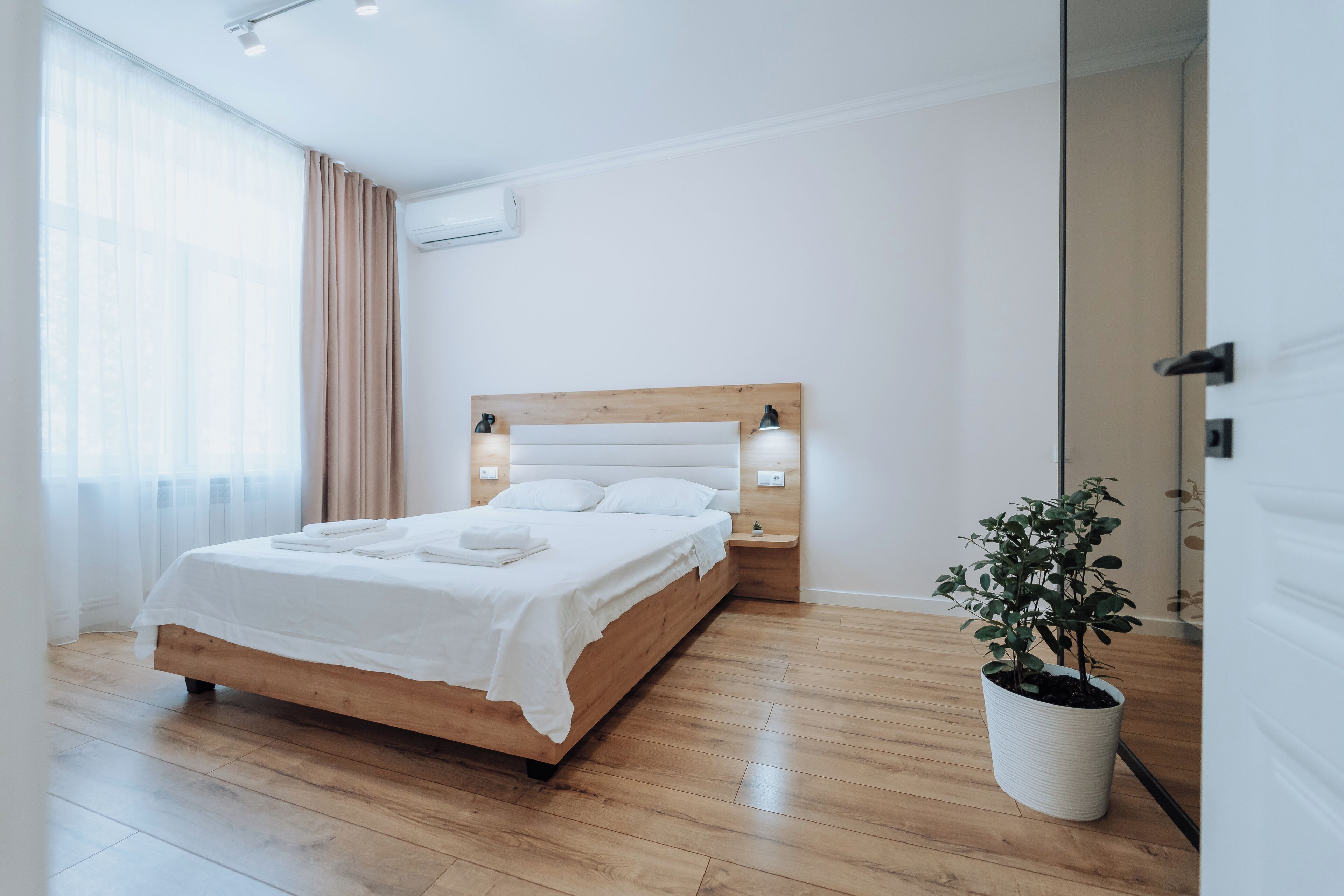 A modern bedroom with a wooden bed, white linens, a bedside table with lamps, and a potted plant.