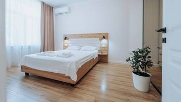 A modern bedroom with a wooden bed, white linens, a bedside table with lamps, and a potted plant.