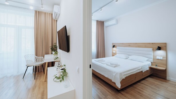 Ein modernes Hotelzimmer mit Holzboden, einem weißen Bett mit Holz Kopfteil, einem Schreibtisch mit Stuhl und einem großen Fenster mit Vorhängen.