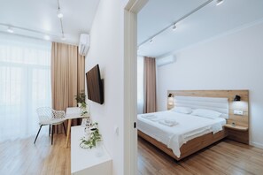 A modern hotel room with a wooden floor, a white bed with a wooden headboard, a desk with a chair, and a large window with curtains.