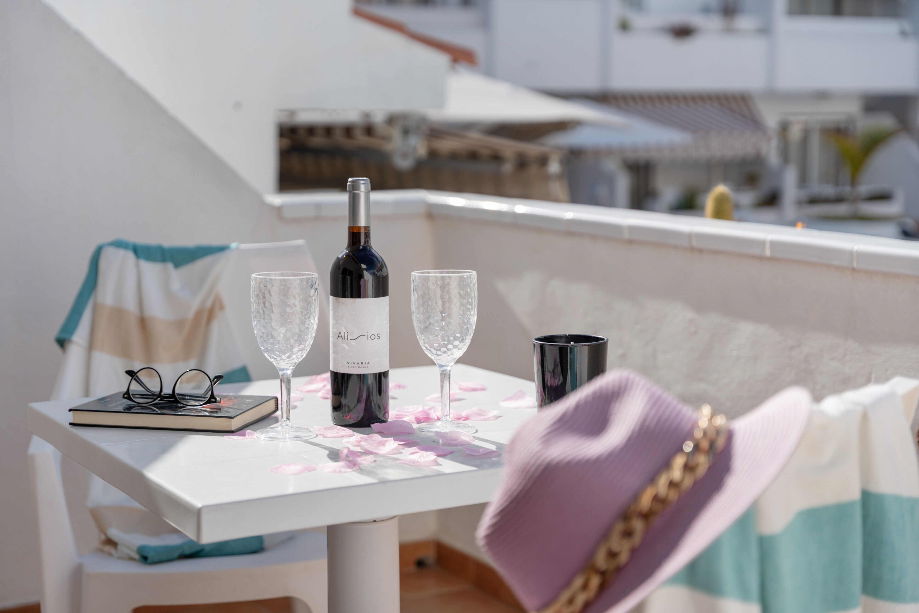 A table with a bottle of wine, two glasses, a notebook, and a pair of glasses on a balcony with a view of buildings.