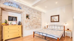 A bedroom with a stone wall, a bed with a patterned bedspread, a wooden dresser, and a small bedside table. - Apartment Irma: Studio apartman (Pula)