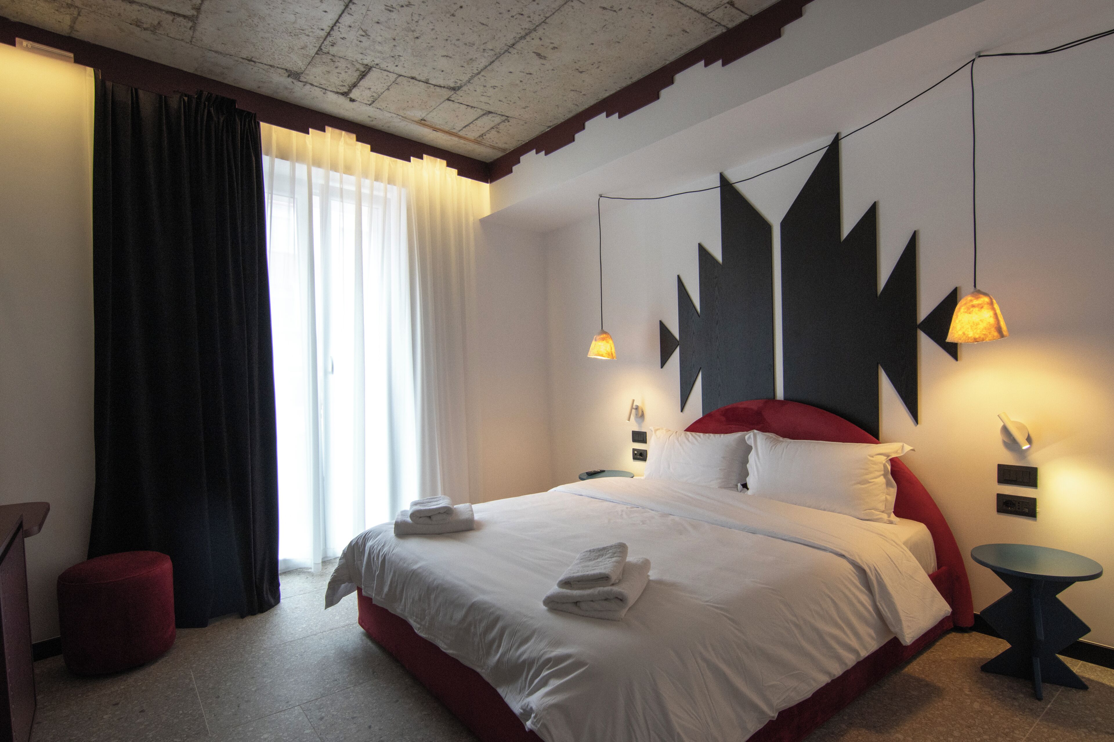 A hotel room with a large bed, a red headboard, a round side table, and a red ottoman.