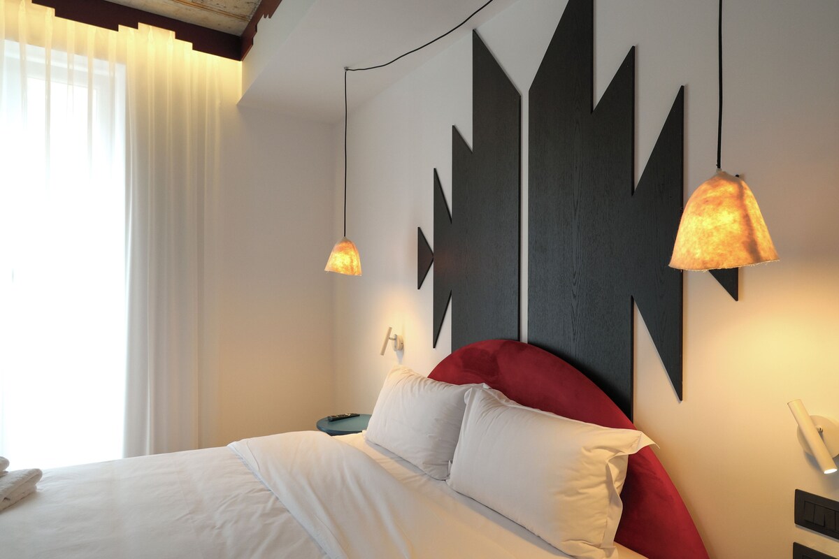 A hotel room with a bed, white bedding, a red headboard, a wall-mounted abstract art piece, and two pendant lights.