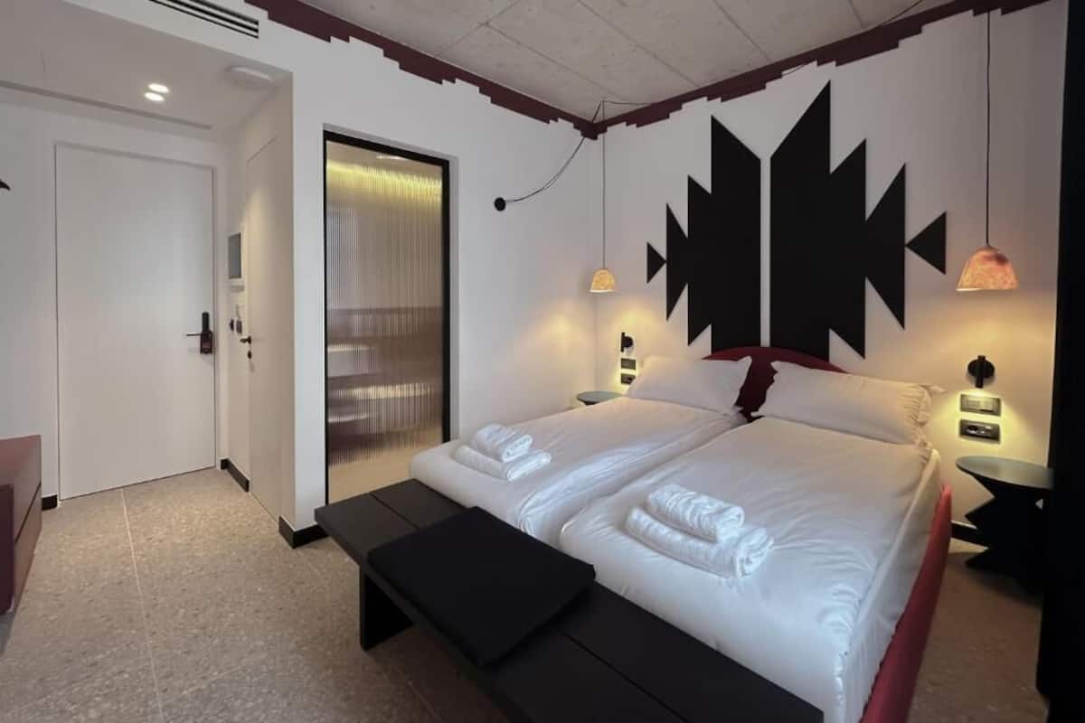A modern hotel room with a large bed, a bench, a door, and a wall with geometric patterns.