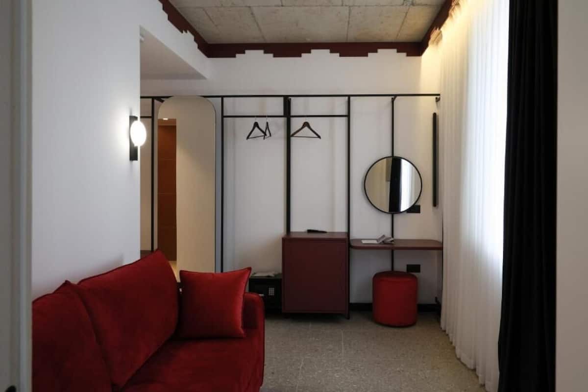 A compact hotel room with a red sofa, a small desk, a mirror, and a wardrobe with hangers.