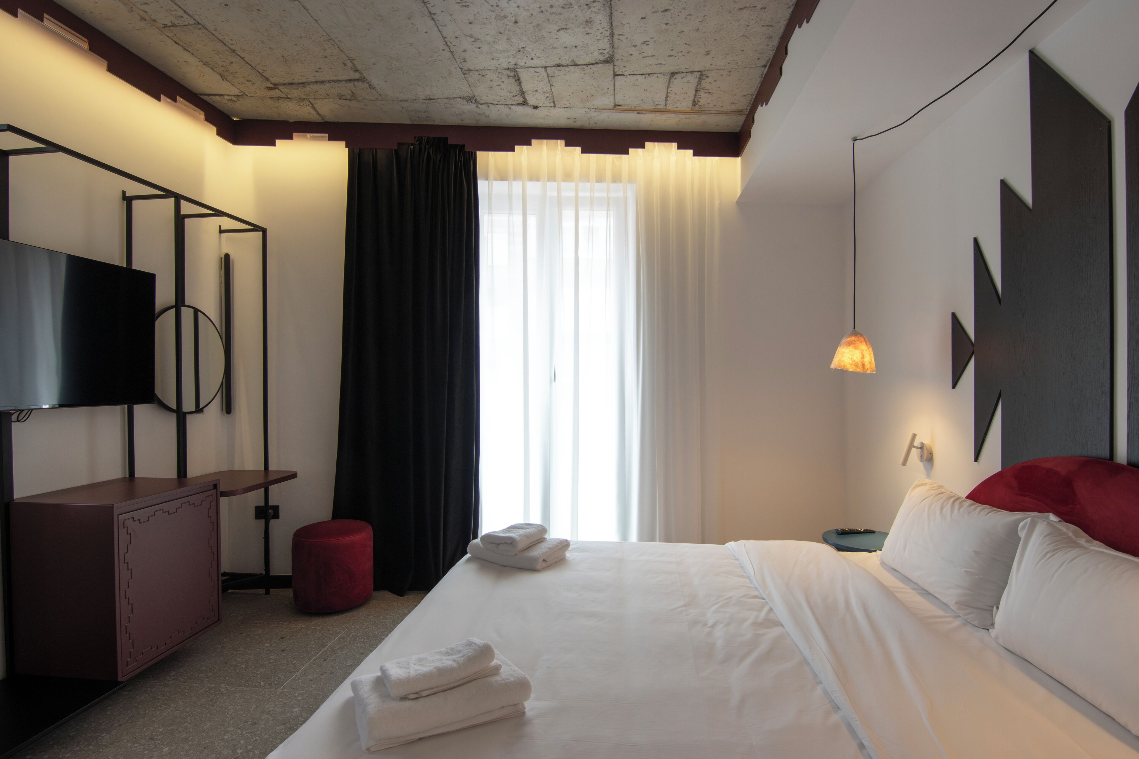 Ein Hotelzimmer mit Bett, Fernseher, Schreibtisch, Hocker in Rot und einem Fenster mit Vorhängen.