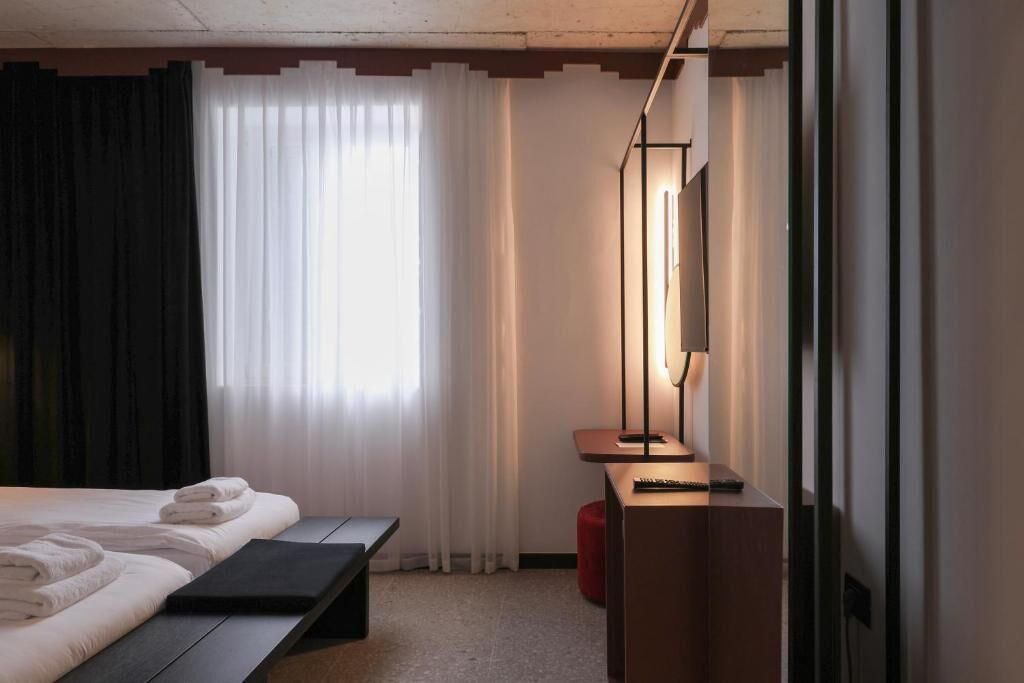 A hotel room with a bed, a bedside table with a lamp, a bench, and a large mirror.