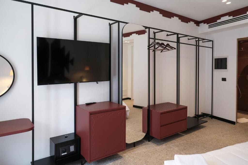 A hotel room with a bed, a TV mounted on the wall, a wardrobe, a small cabinet, and a hanging clothes rack.