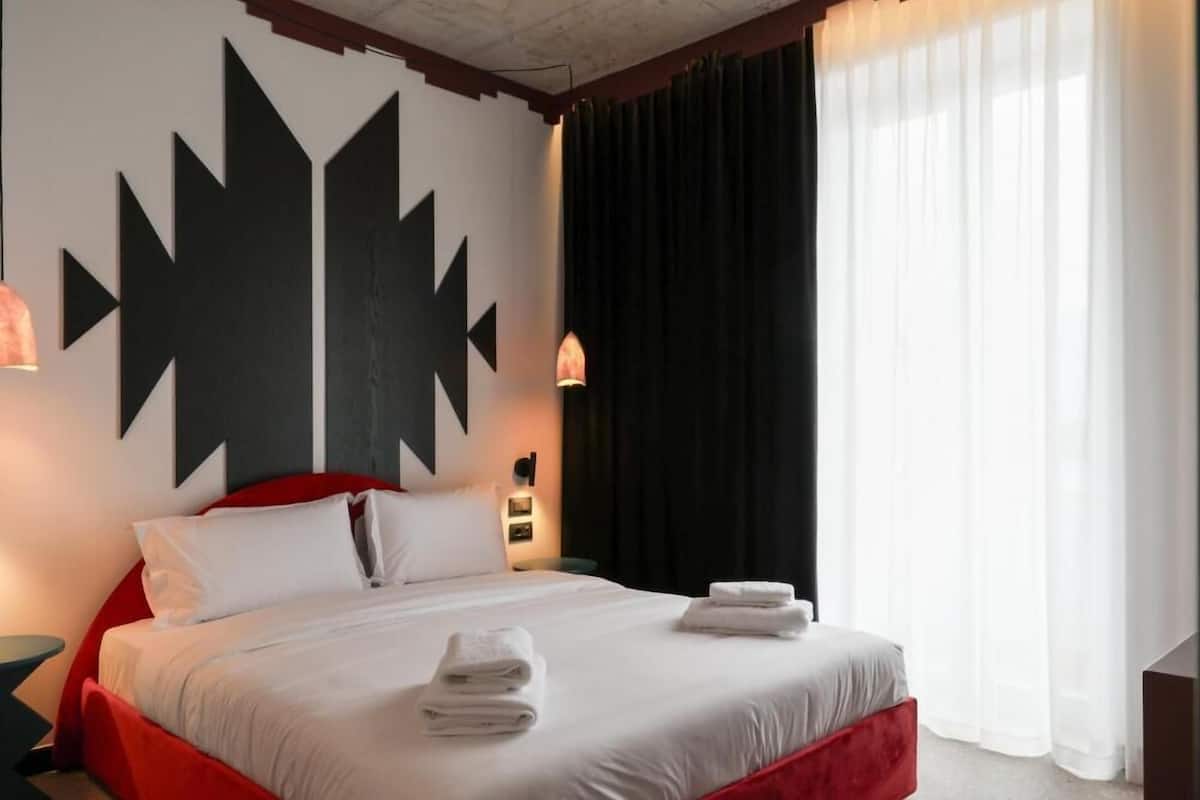 A hotel room with a large bed, white bedding, red accents, and a modern wall design.