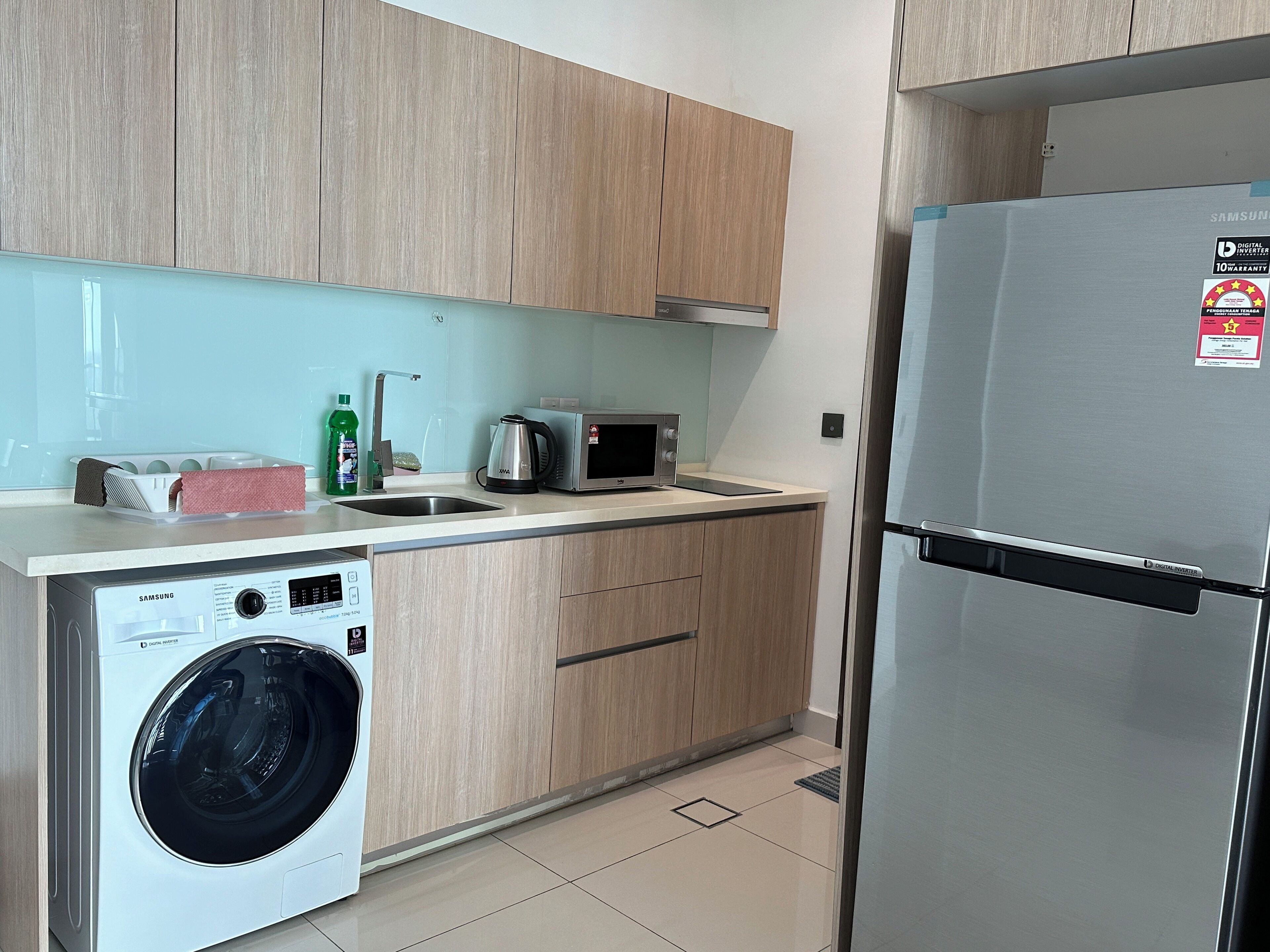 A modern kitchen with a stainless steel refrigerator, a microwave, a washing machine, and wooden cabinets.