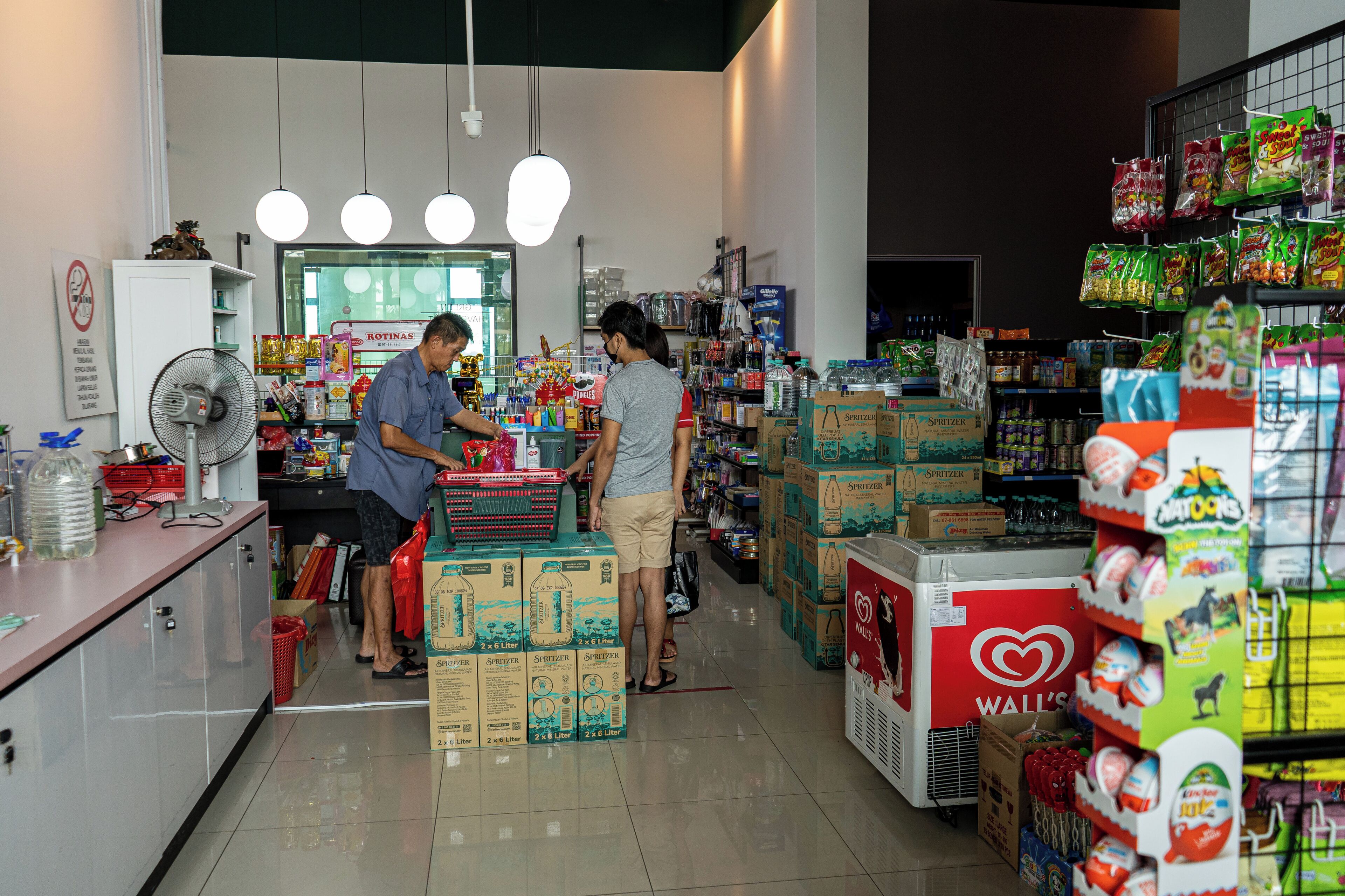 A grocery store interior with shelves stocked with various products, a refrigerated display, and a fan on the counter.