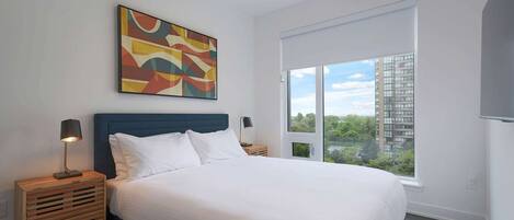 A modern bedroom with a large bed, a colorful abstract painting, a nightstand with a lamp, and a window with a view of buildings and trees.