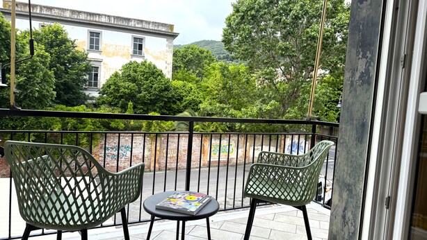 Varanda com duas cadeiras verdes e uma pequena mesa, com vista para uma rua com grafites na parede e árvores ao fundo.