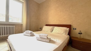 Cama bem arrumada com lençóis e toalhas brancas, cabeceira de madeira, mesa de cabeceira com abajur e vista da cidade pela janela com cortinas.