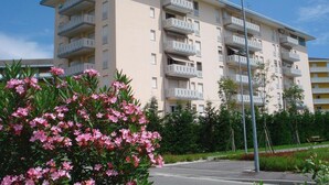 Un edificio residencial de varios pisos con balcones, un área pavimentada en la parte delantera y vegetación circundante.
