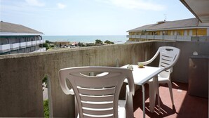 Un balcón con dos sillas de plástico blanco y una mesa, con vista a una playa y un edificio de fachada amarilla y blanca.