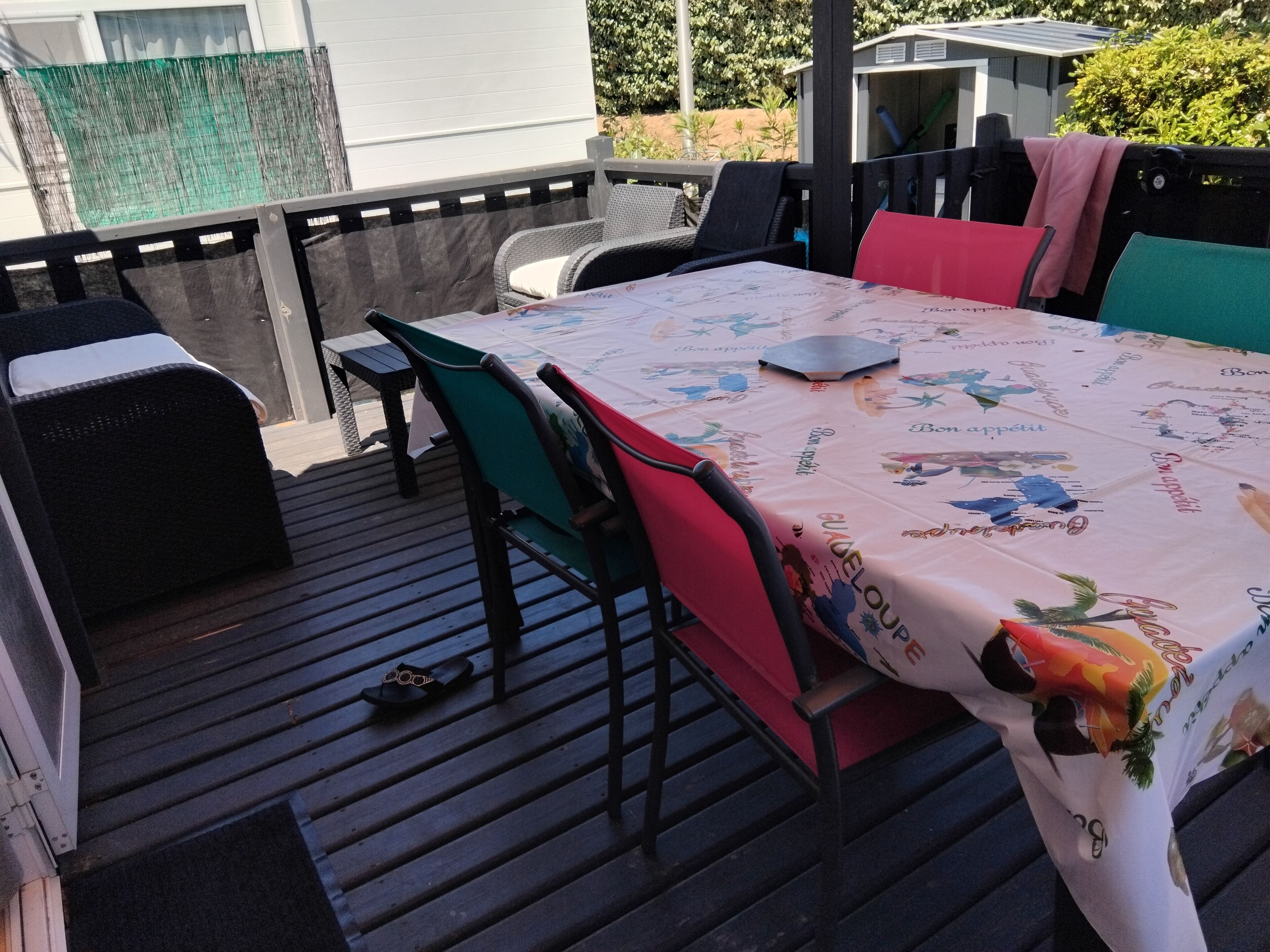 En udendørs patio med et bord dækket af en dug med tekst og billeder, omgivet af fletstole og en bænk.