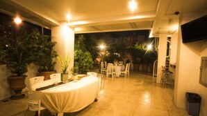 Un espace extérieur abrité, aménagé pour prendre les repas, avec une table et des chaises, entouré de plantes en pot et d’un téléviseur fixé au mur.