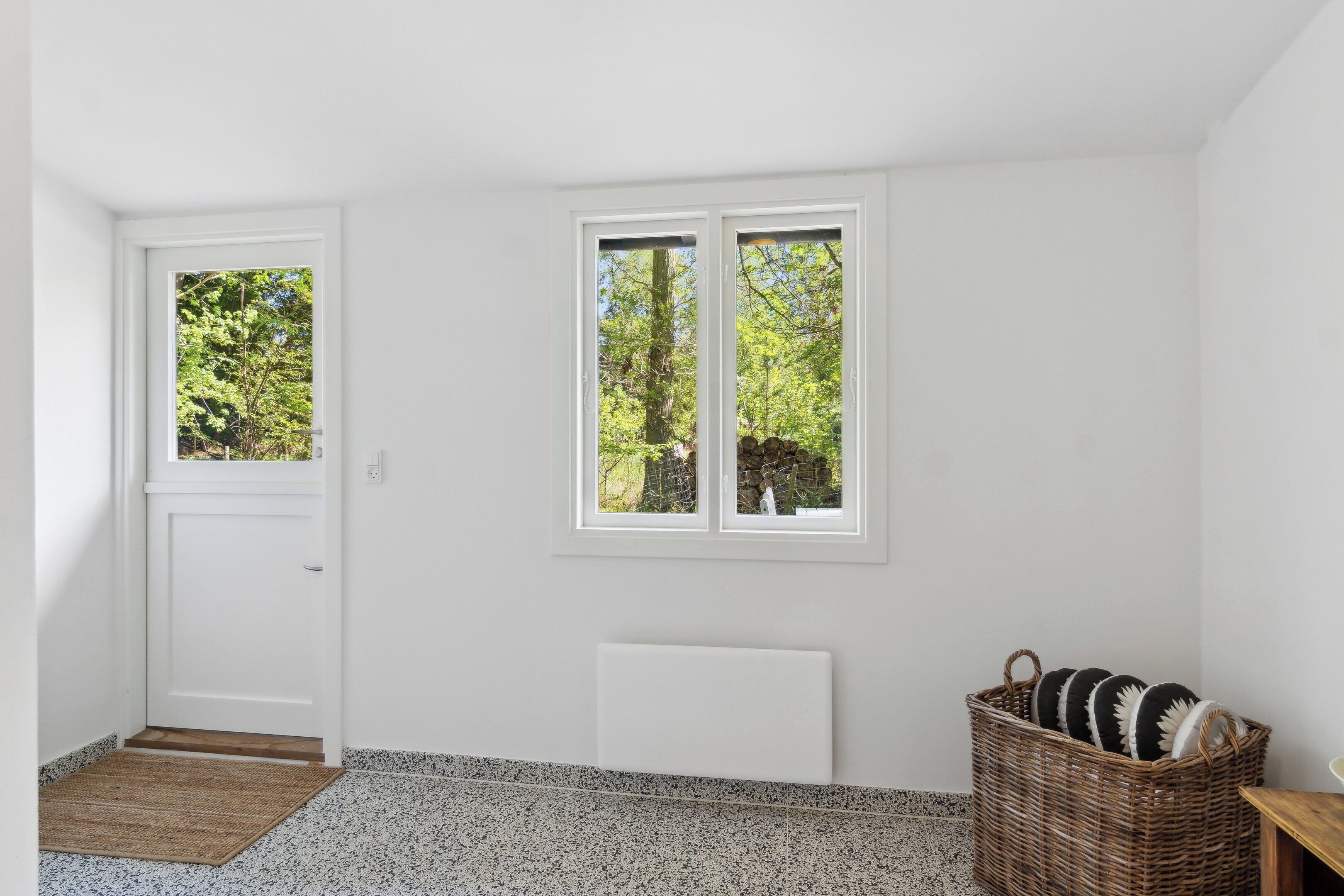 Ein Zimmer mit weißer Tür, zwei Fenstern, einer Heizung, einem Korbwägelchen und einer Holzbank.