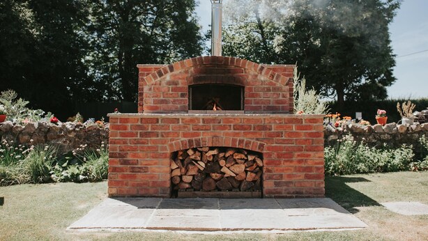 Un horno de pizza de ladrillo al aire libre con chimenea, rodeado por un muro de piedra y un jardín.