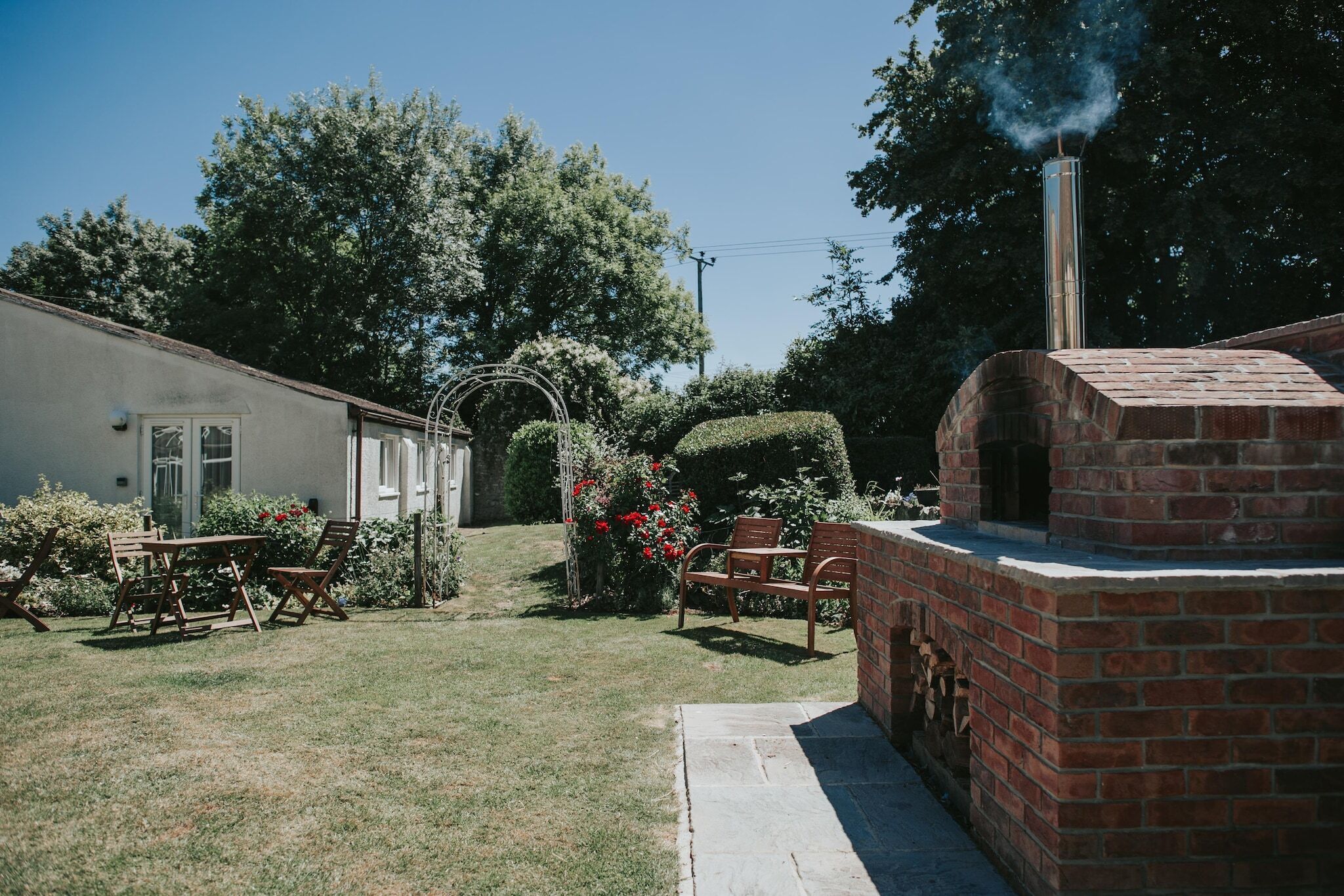 Un four extérieur en briques avec conduit de fumée, une terrasse avec des meubles en bois et un jardin agrémenté de fleurs et d’une haie.
