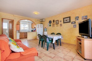 Un salon avec un canapé, une table à manger, des chaises, un téléviseur et, au fond, un coin cuisine.