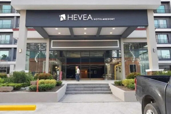 Une entrée d’hôtel moderne avec une façade en verre, un espace d’entrée abrité et un jardin paysager à l’avant.