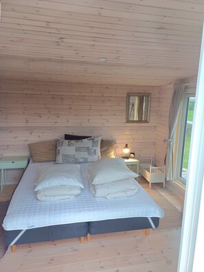 Een slaapkamer in een houten chalet, met een bed, kussens, een nachtkastje, een lamp en een raam.