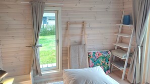 A wooden room with a bed, a window with curtains, a shelf with folded towels, and a view of a house outside. - Lovely cottage close to the water (Karrebæksminde)