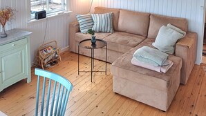 A room with a wooden floor, a beige sofa, a small round table, a blue chair, a window with a view, and a shelf with books and a plant. - Lovely cottage close to the water (Karrebæksminde)