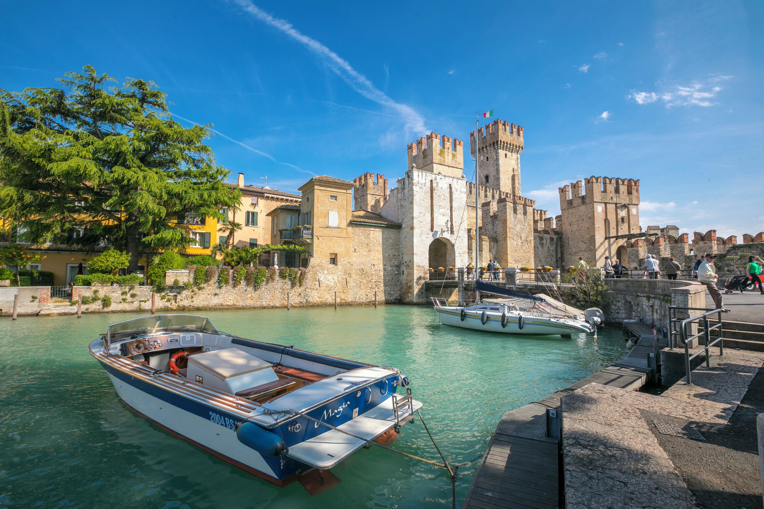 Un castillo histórico con un foso, barcos amarrados en el muelle y un cielo azul despejado.