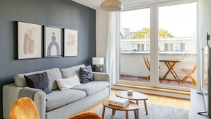 A modern living room with a grey sofa, a small wooden table, and a large window offering a view of the city. - Blueground | Josefstadt, terrace, nr Votivkirche (Vienna)