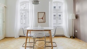 A modern room with a wooden herringbone floor, a white table with transparent chairs, a pendant light, and a floor lamp. - Blueground | Alsergrund, view, nr Votivkirche (Vienna)