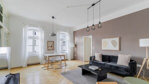 A spacious living area with a dark sofa, a coffee table, and a dining table with chairs. There are large windows with white curtains, a standing lamp, and a framed picture on the wall. - Blueground | Alsergrund, view, nr Votivkirche (Vienna)