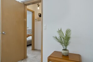 Un couloir avec une porte en bois, une petite table en bois sur laquelle est posée une plante en pot, et un lit à l’arrière-plan.