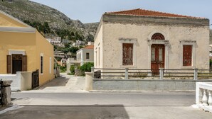 Une rue bordée d’immeubles, un immeuble jaune sur la gauche, un immeuble blanc avec un toit recouvert de tuiles rouges au centre, et une montagne en arrière-plan.