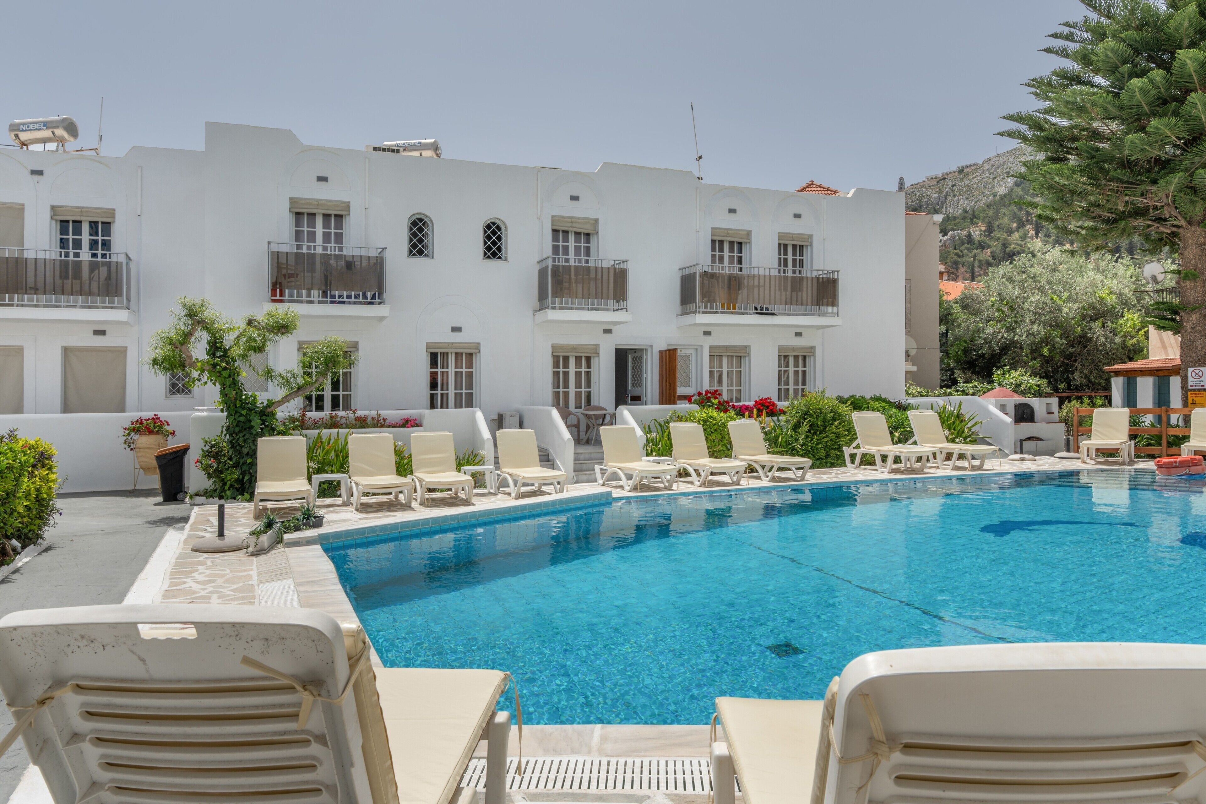Una piscina con sillones de descanso, un edificio blanco con balcones y una montaña al fondo.