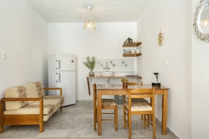 Un espace de vie compact comprenant un canapé, une table à manger, des chaises, un réfrigérateur, un coin cuisine avec un évier et un plan de travail, ainsi qu’un luminaire suspendu.