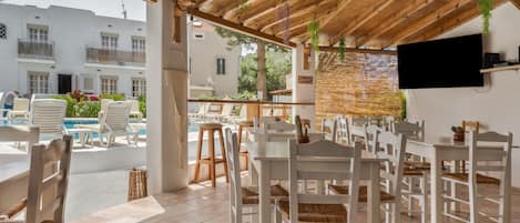 A covered outdoor seating area with white tables and chairs, a pool, and a TV mounted on the wall.
